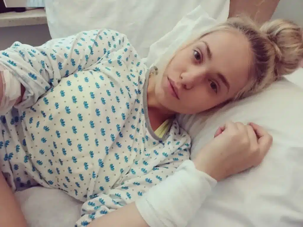 Janita Lukkarinen makaa sairaalasängyssä potilasvaatteissa katsoen kameraan rauhallisella mutta väsyneellä ilmeellä syöpädiagnoosin jälkeen vuonna 2018